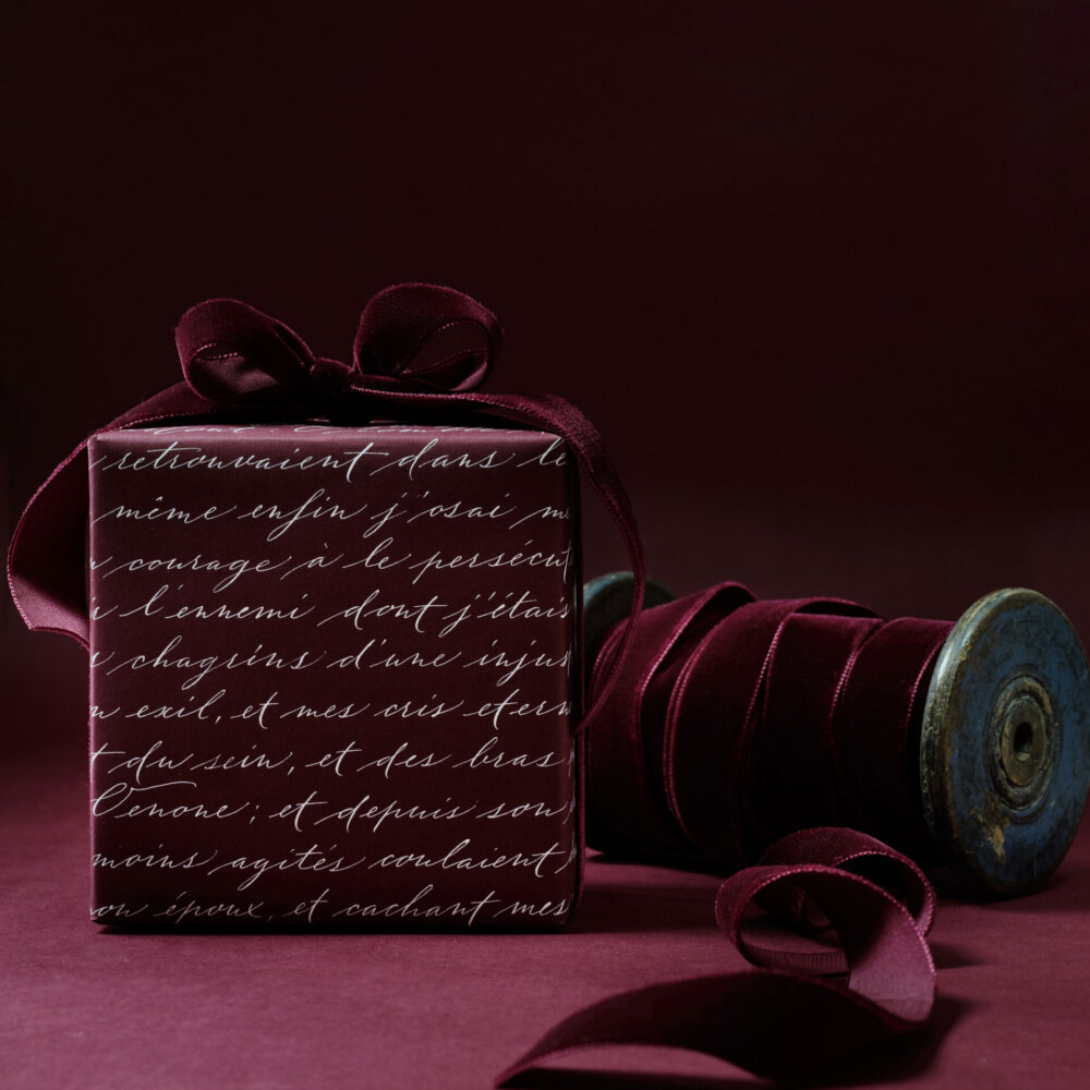 Un cadeau luxueux enveloppé dans du papier bordeaux profond orné d'un élégant script manuscrit. L'emballage est noué avec un ruban en velours doux, complétant parfaitement la palette de couleurs riche. Un rouleau de ruban en velours assorti repose à proximité, renforçant l'atmosphère sophistiquée et raffinée de la scène.